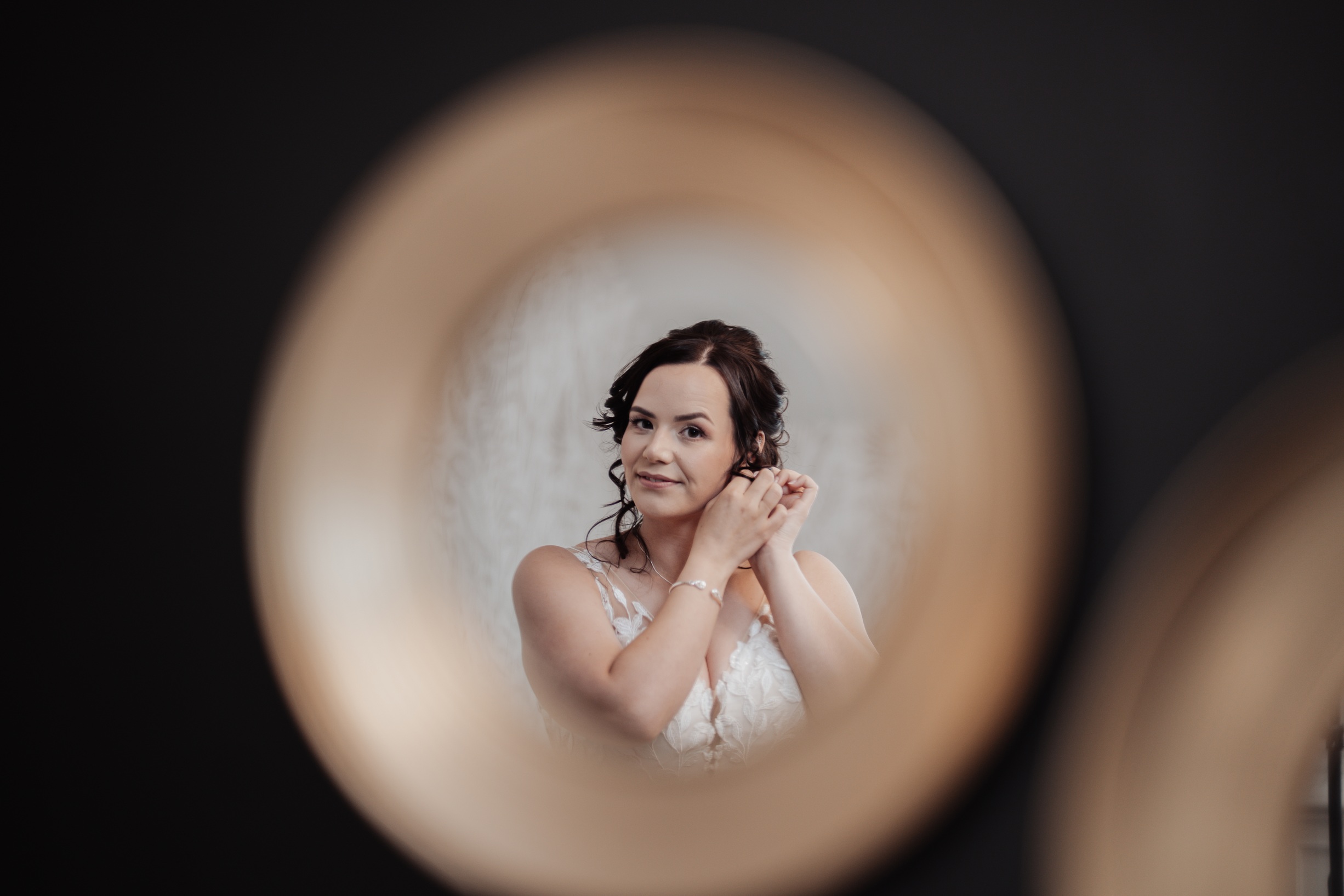 A reflective close-up photo of a bride in her wedding dress, seen through a circular mirror, as she adjusts her earring. 
                        The photo captures her smiling expression as she prepares for her special day at Llanerch Vineyard.
