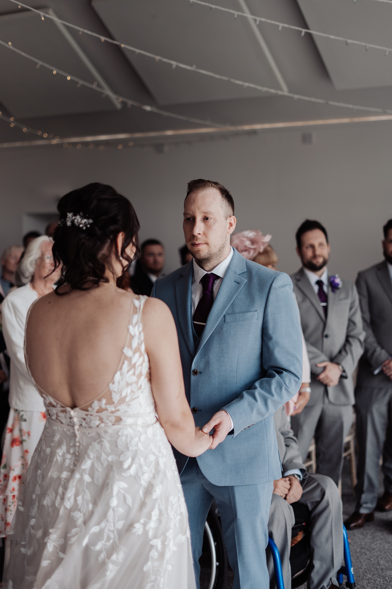 A candid shot of a bride and groom holding hands during their wedding ceremony at Llanerch Vineyard. They are looking at each other, 
                        and guests are visible in the background.