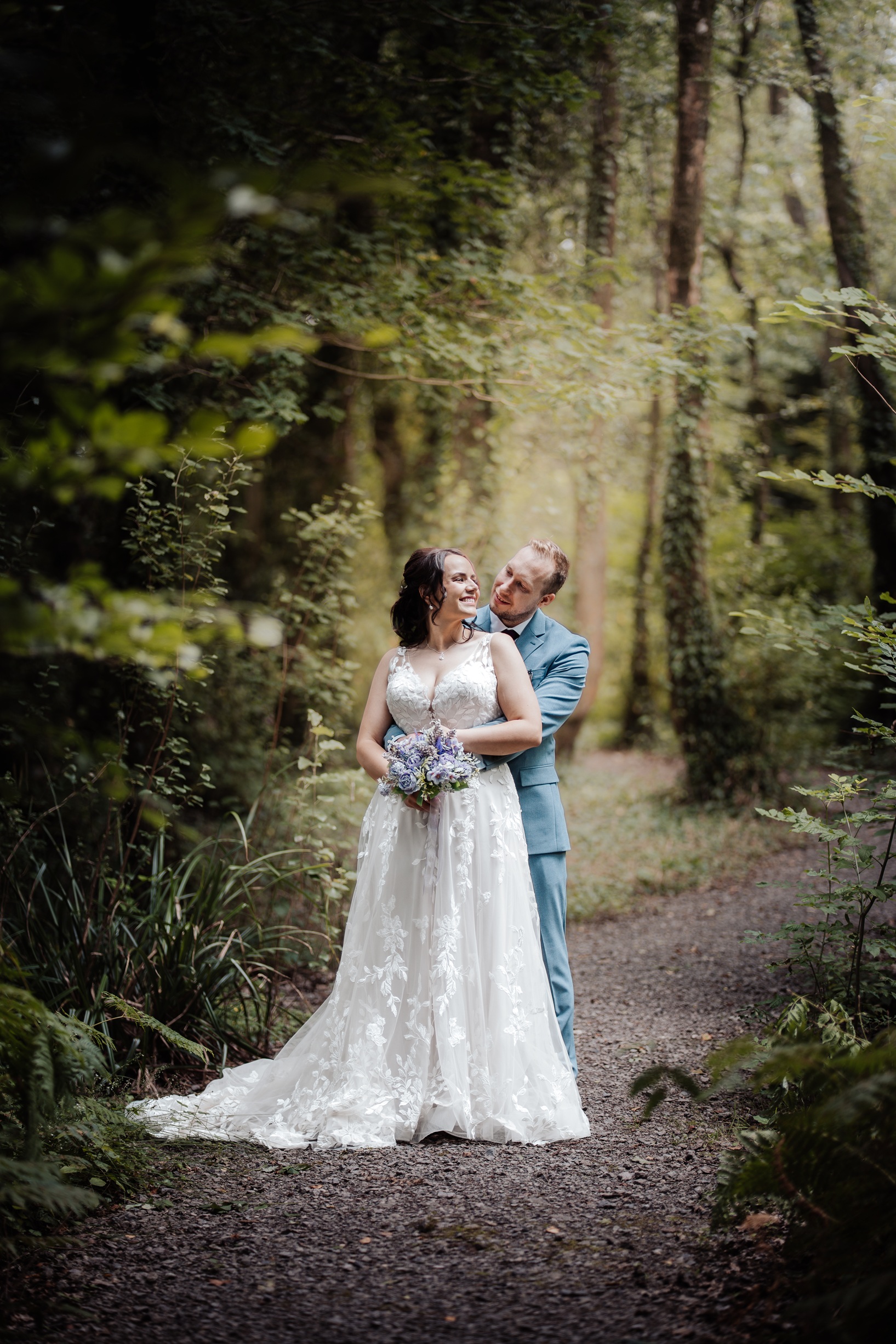 The groom embraces his bride from behind while they pose on a wooded path, both smiling. The background is a mix of green leaves and trees, creating a beautiful 
                        setting for their wedding photos.