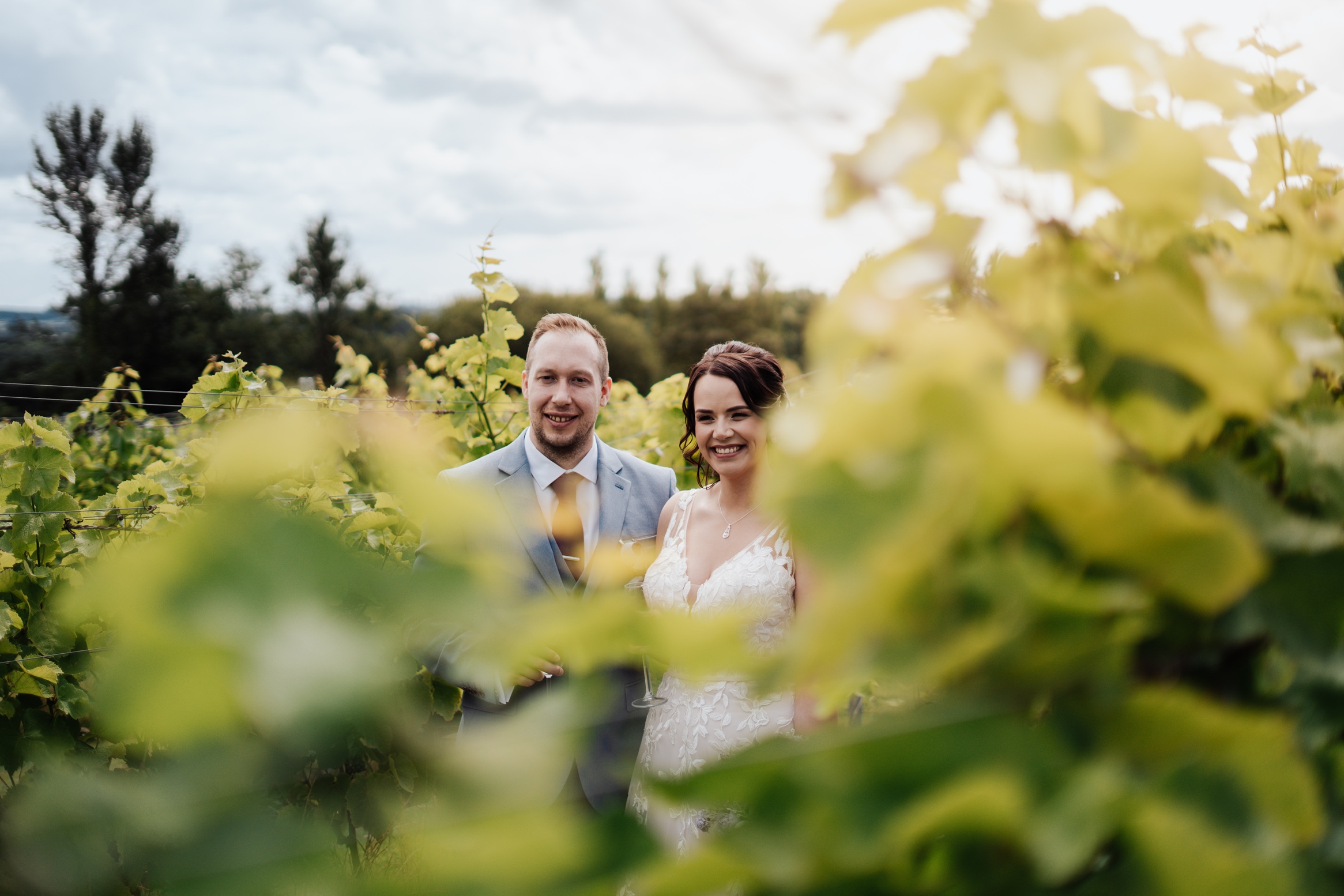 A bride and groom pose together in a vineyard, with golden leaves framing the shot. They are both smiling, holding a glass of wine, and looking at the camera.