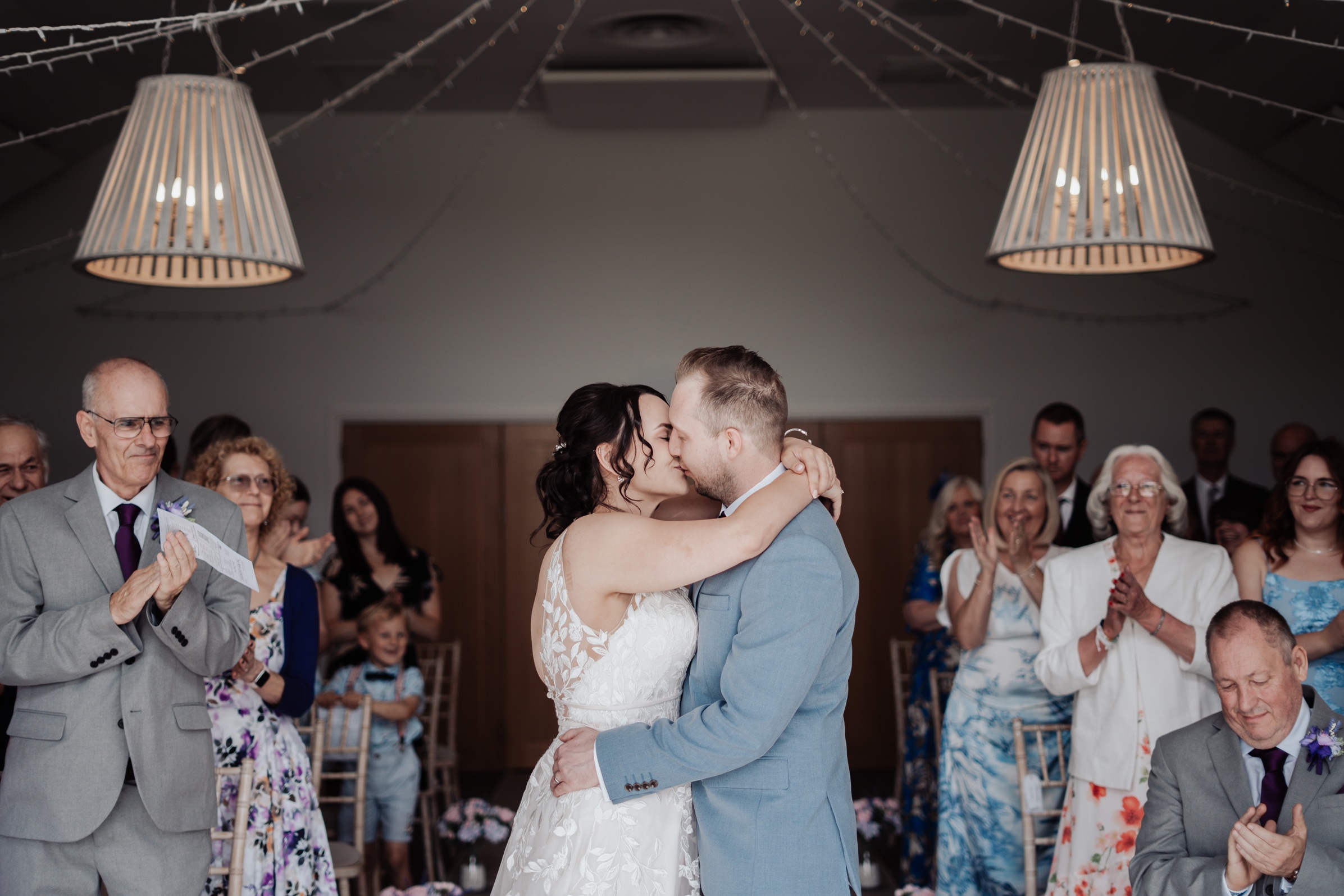 The newly married couple shares a kiss at the end of their wedding ceremony, with happy guests clapping in the background at Llanerch Vineyard, 
                        South Wales. The room is decorated with draped fabric and hanging lights.