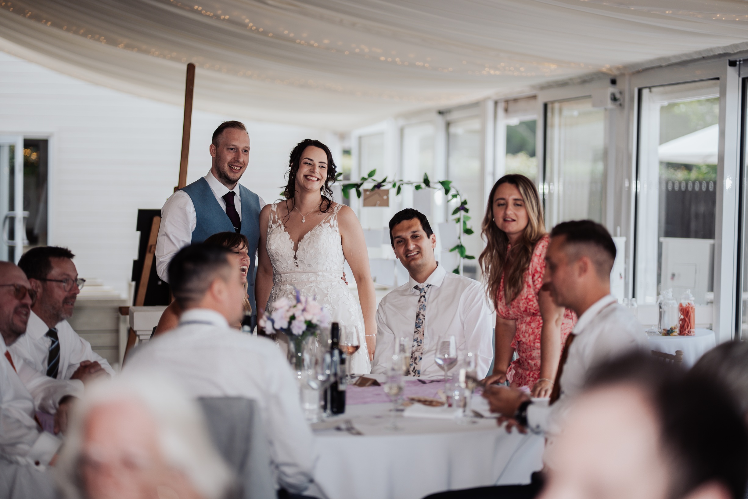 A candid photo of a bride and groom laughing with their wedding guests while seated at a reception table at Llanerch Vineyard, South Wales. 
                        The room is decorated with a white-draped ceiling and fairy lights.