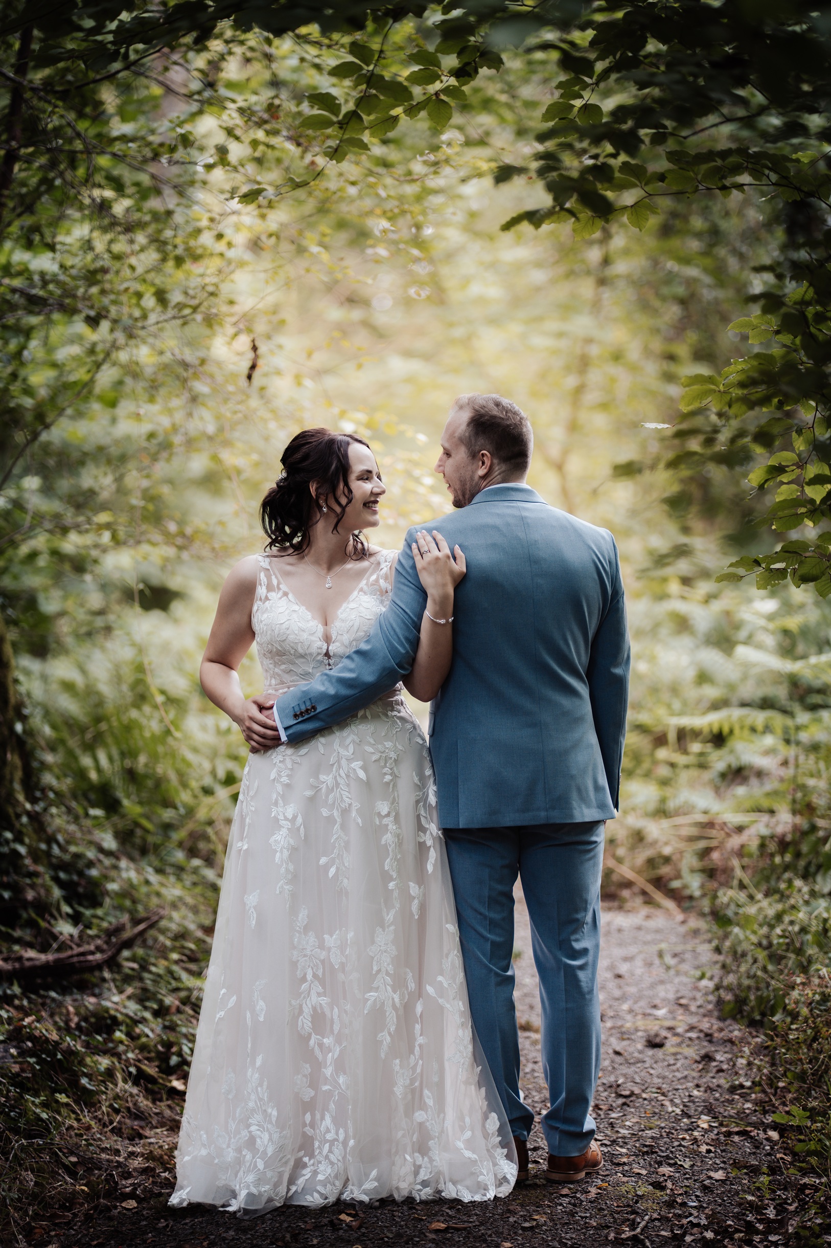 A bride and groom look back over their shoulders at the camera while walking away from the viewer on a wooded path. The groom has his arm around the bride.