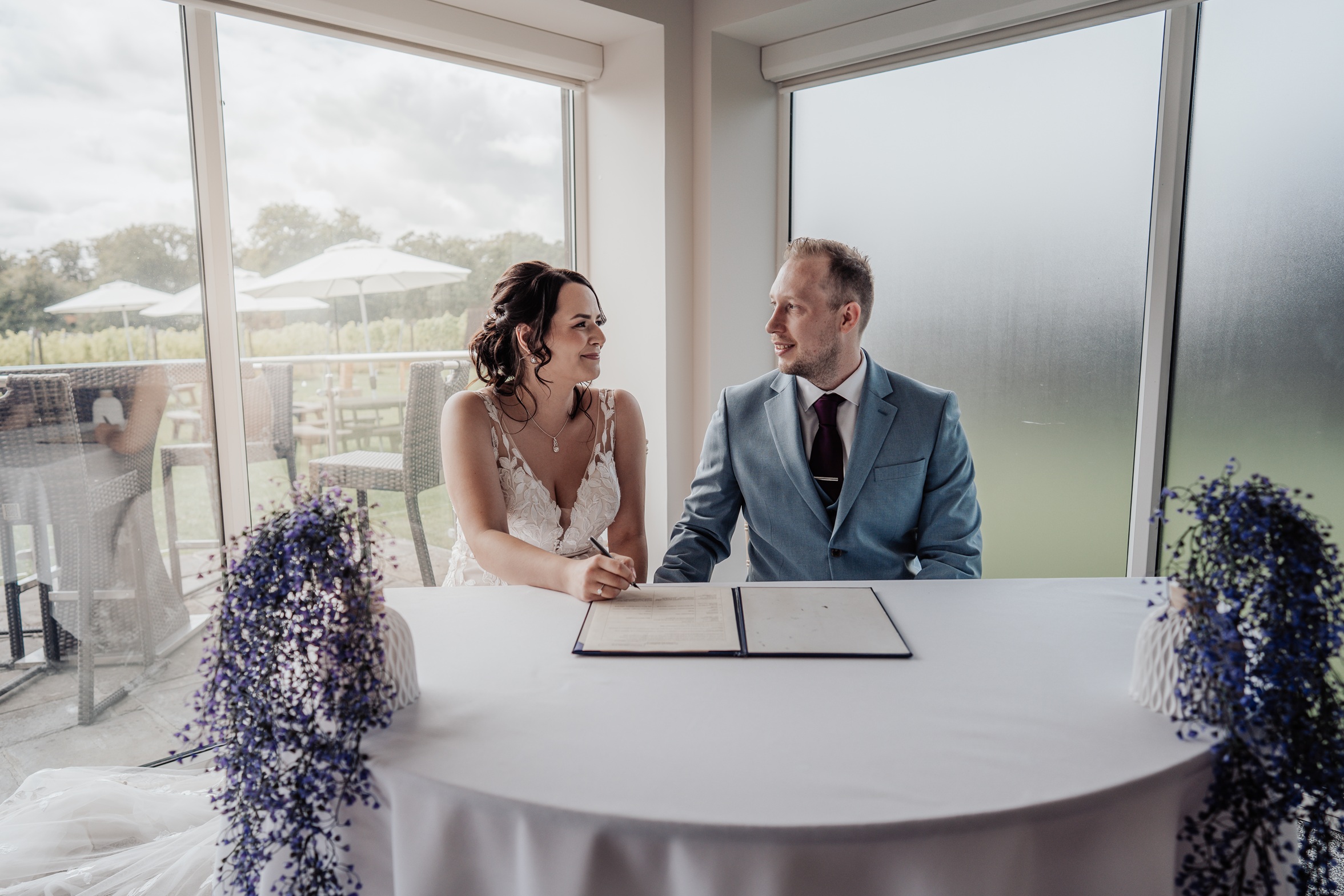 A couple in their wedding attire signing their marriage certificate. They are seated at a table near a large window, looking at each other and smiling.