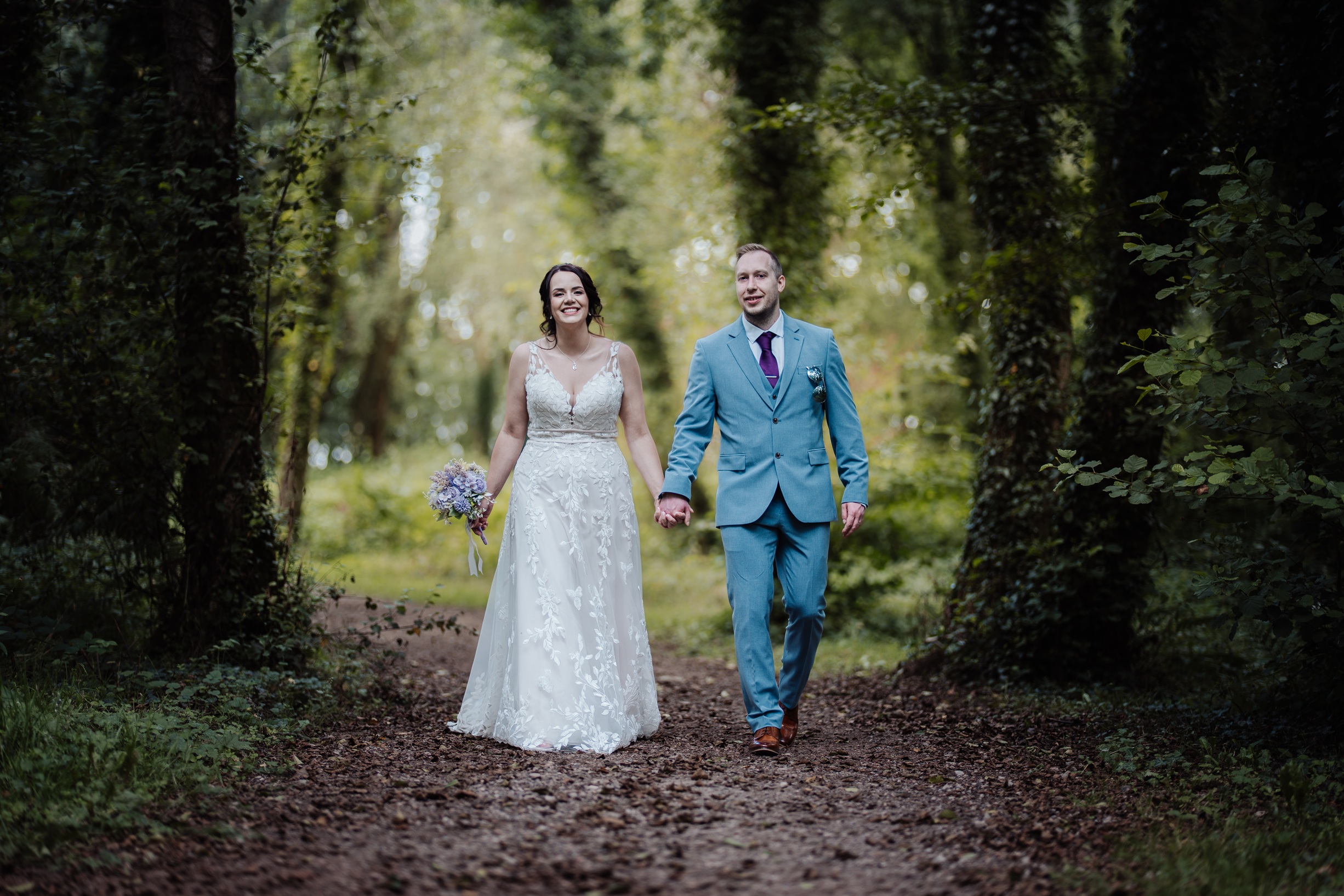 A happy bride and groom walk hand-in-hand down a wooded path, surrounded by lush green foliage.
                        The photo captures a romantic moment from their wedding day at Llanerch Vineyard, South Wales.