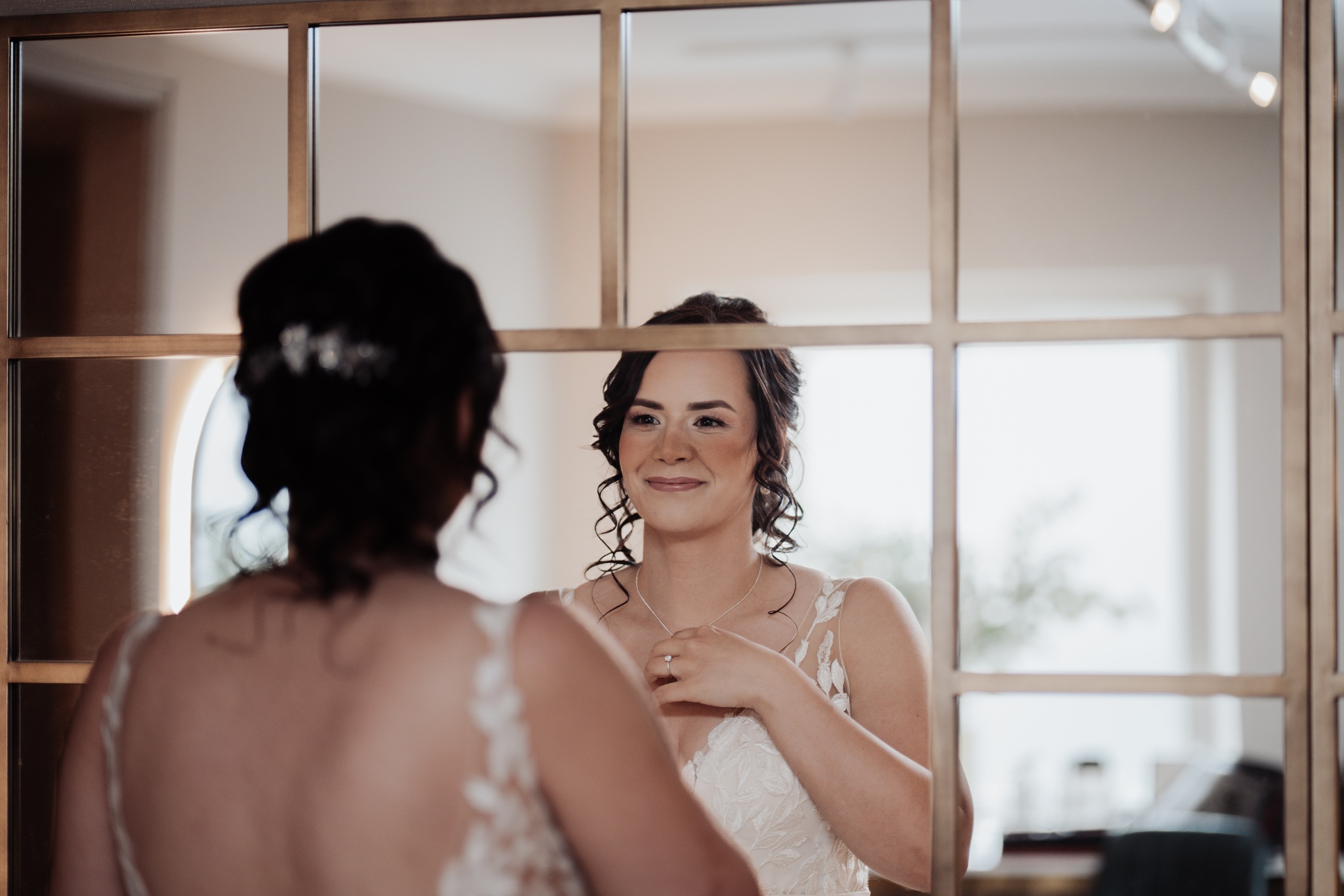 A reflective photo of a bride smiling at her own reflection in a large, gridded mirror while getting ready for her wedding at 
                        Llanerch Vineyard in South Wales. She is wearing a lace wedding dress and a necklace.