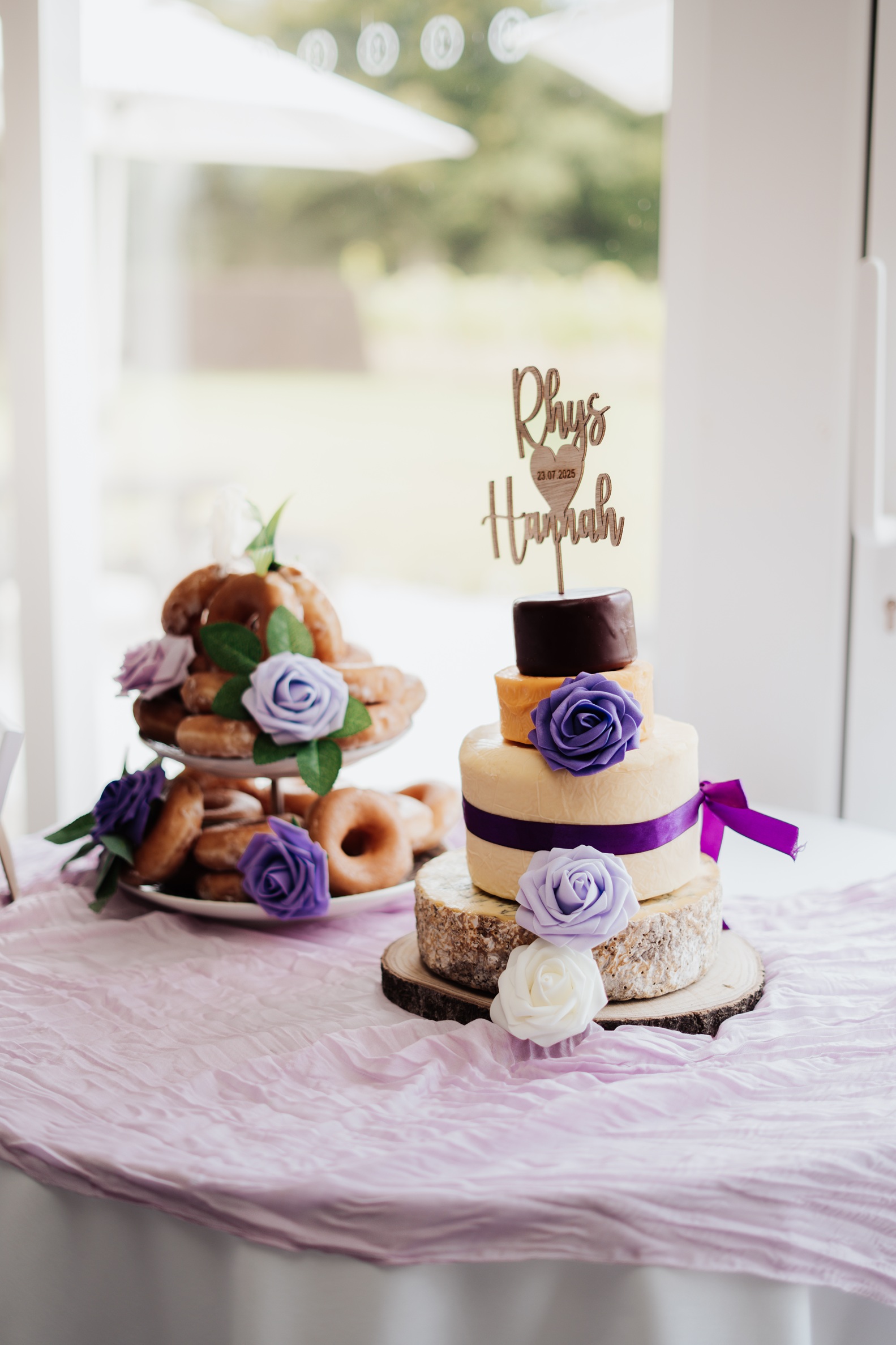 A close-up photo of a wedding 'cake' made of stacked cheese wheels, decorated with purple flowers and a custom topper with the names 'Rhys & Hannah.' In the background, a tiered stand holds donuts.