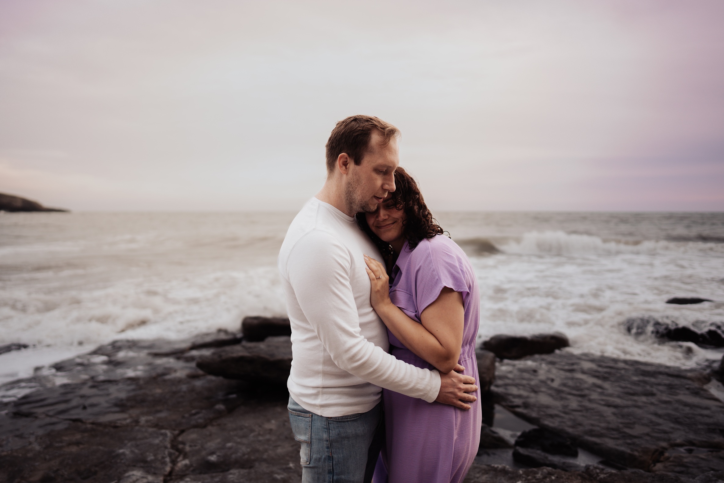 A couple in a warm embrace, standing on a grassy clifftop overlooking the sea and the ruins of Dunraven Castle at Dunraven Bay, Wales. 
                        The golden hour light creates a beautiful, romantic atmosphere.
