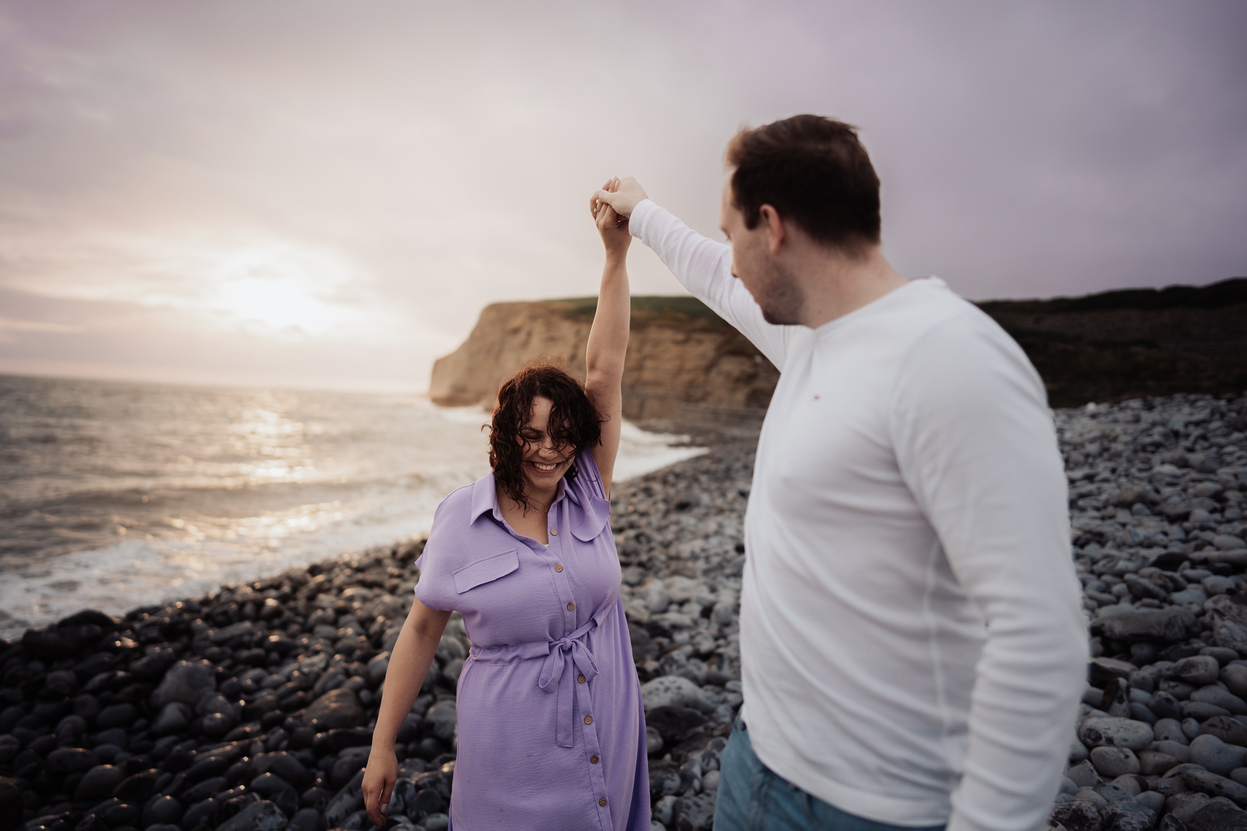 A happy couple walks hand-in-hand along the sandy beach at Dunraven Bay, South Wales, with a backdrop of dramatic cliffs and blue sky.