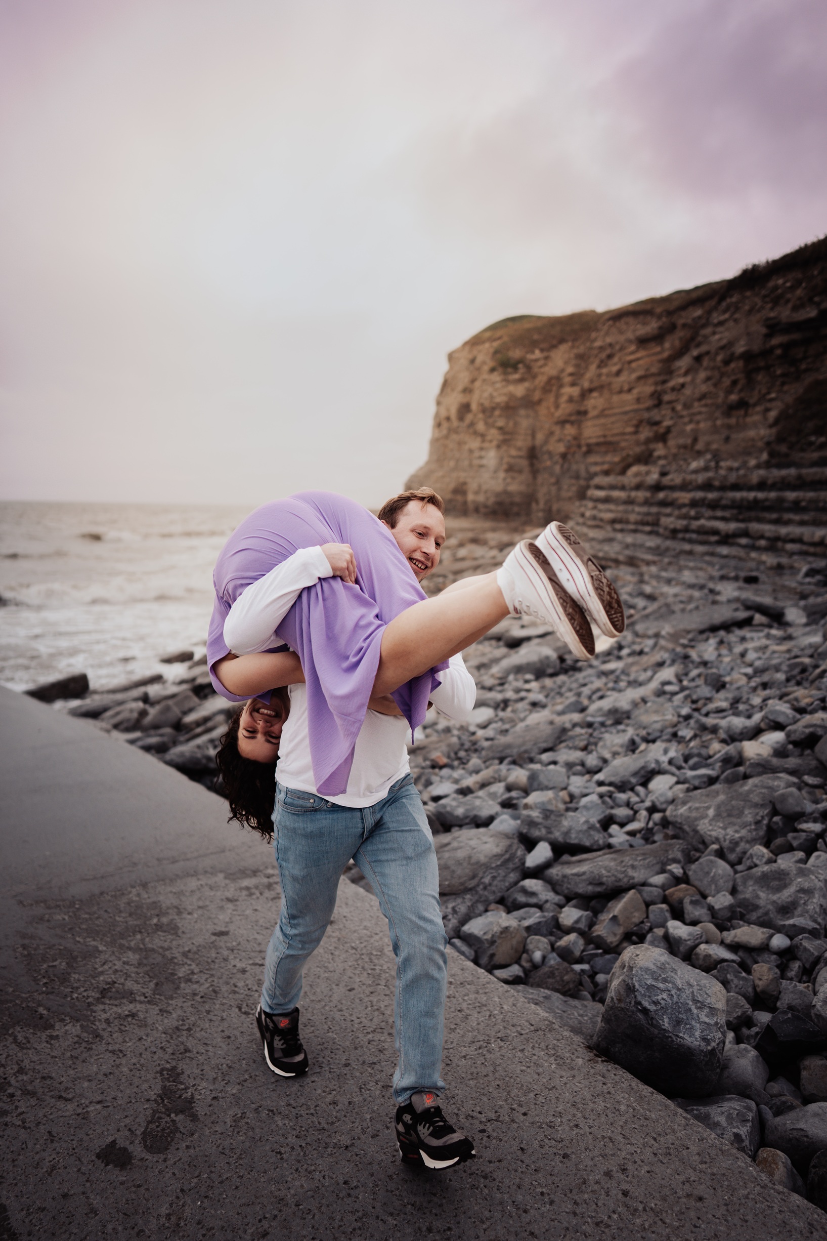 An engaged couple holding hands and walking up a grassy path on a cliffside, with the expansive 
                        sea visible behind them at Dunraven Bay, South Wales.