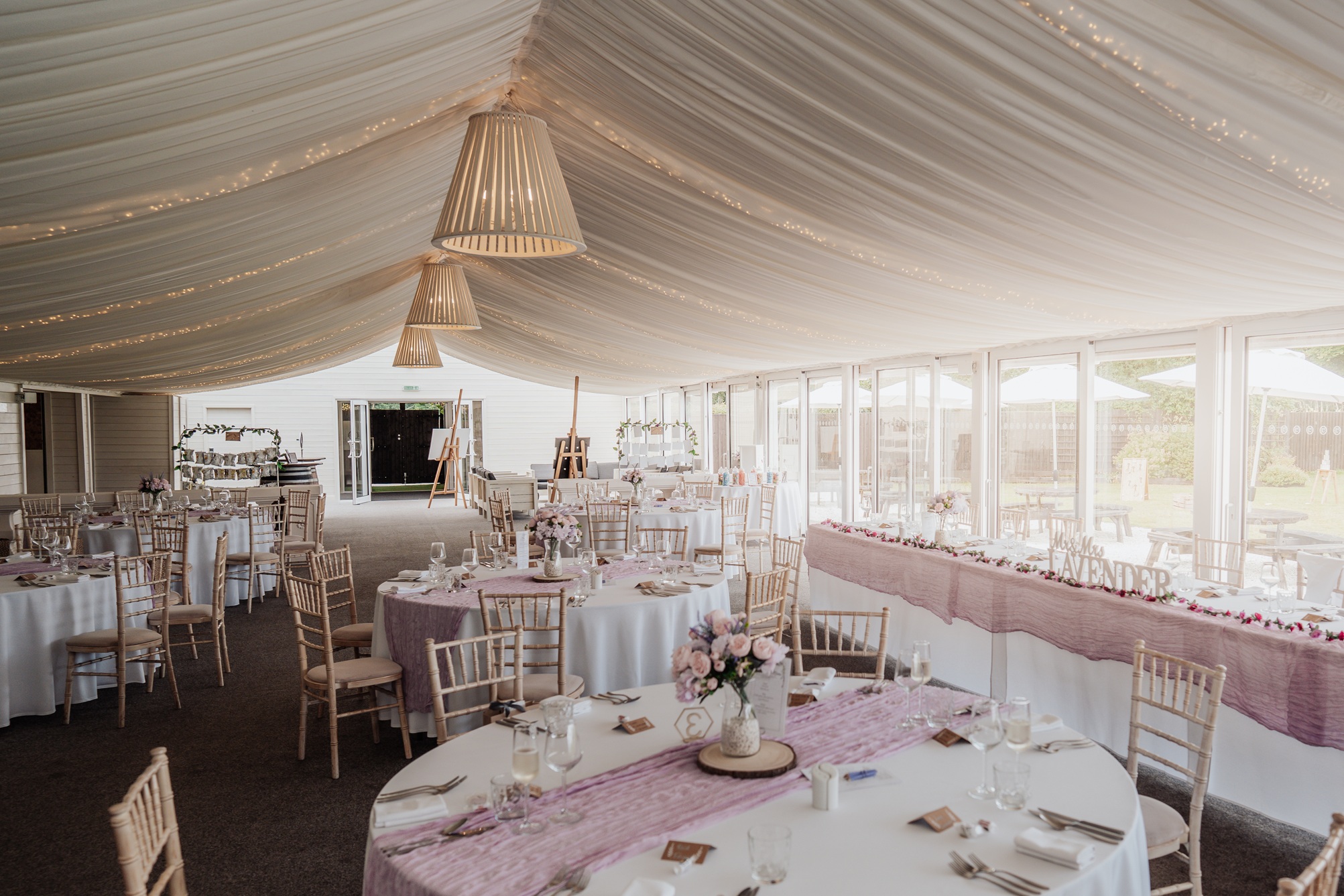 A wide shot of the empty wedding reception room at Llanerch Vineyard, with round tables decorated with pink runners and flowers, ready for the guests. 
                        The room has large windows and a white draped ceiling with fairy lights.