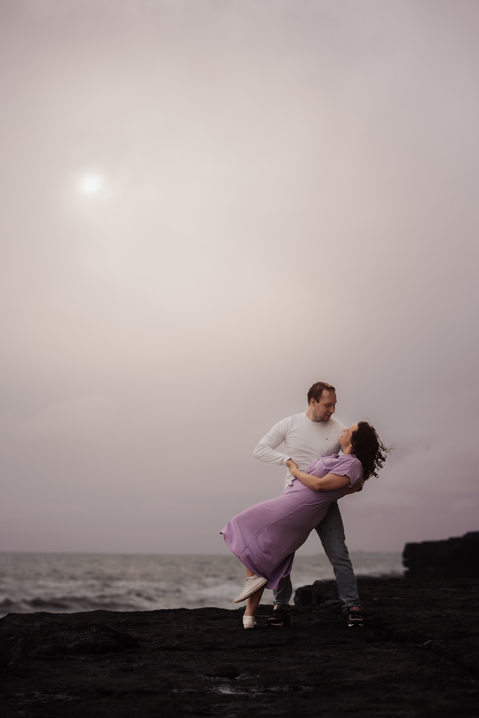 A silhouette of an engaged couple standing together on a clifftop at sunset. The vibrant colors of the setting sun cast a 
                        beautiful glow over the sky and the sea at Dunraven Bay, Wales.