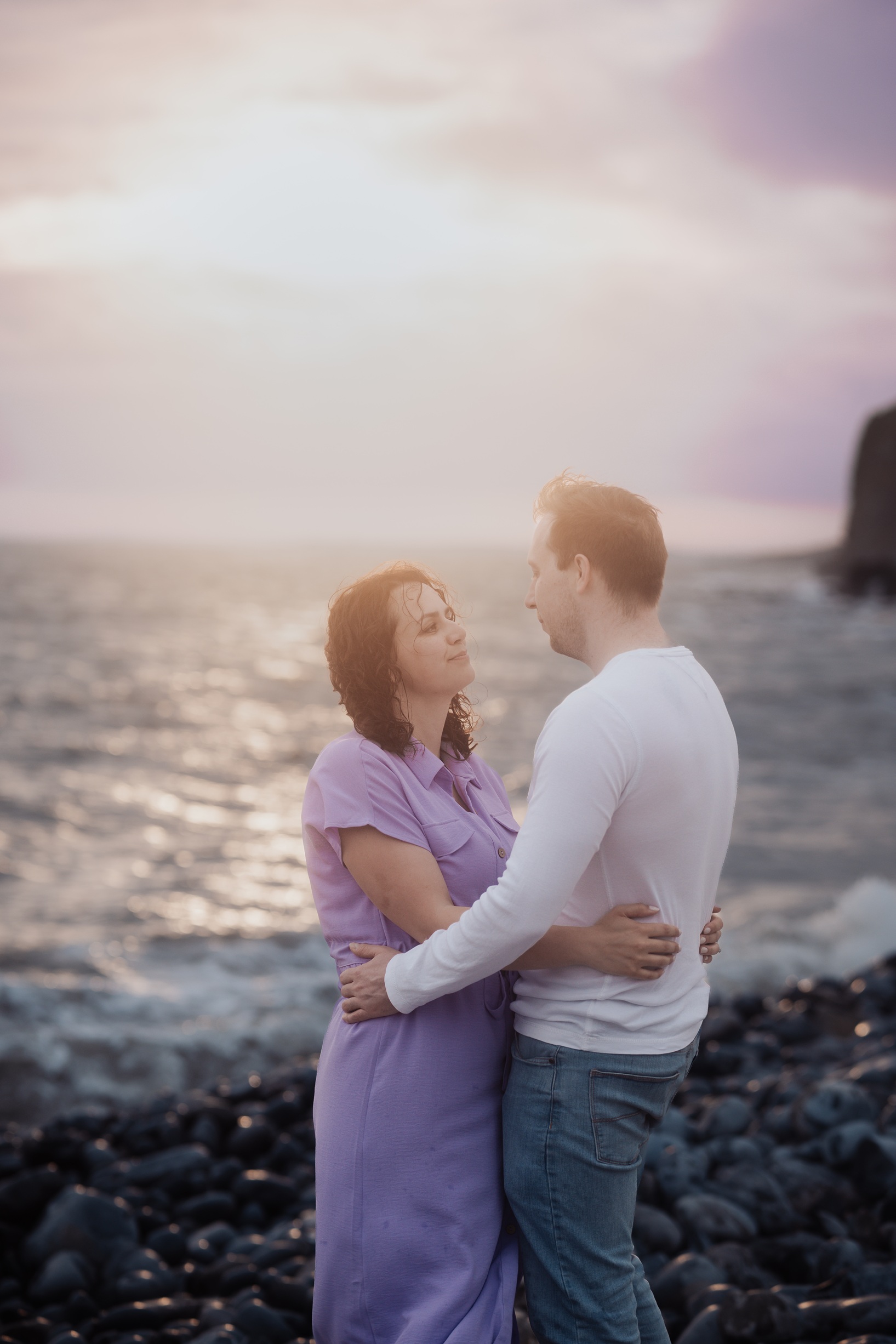 A candid, close-up photo of an engaged couple standing on the rocks at Dunraven Bay, South Wales. 
                        They are smiling at each other with the sea and sky in the background.