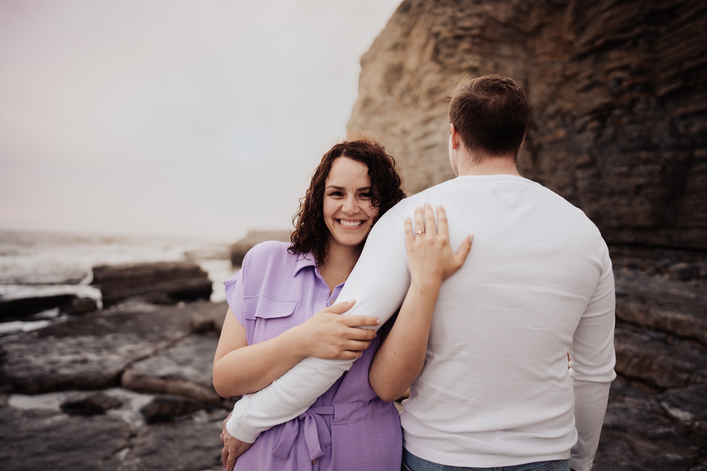 A portrait of an engaged couple standing on a rocky path with dramatic cliffs and the sea in the background. 
                        The photo captures a romantic moment from their photoshoot at Dunraven Bay, Wales.