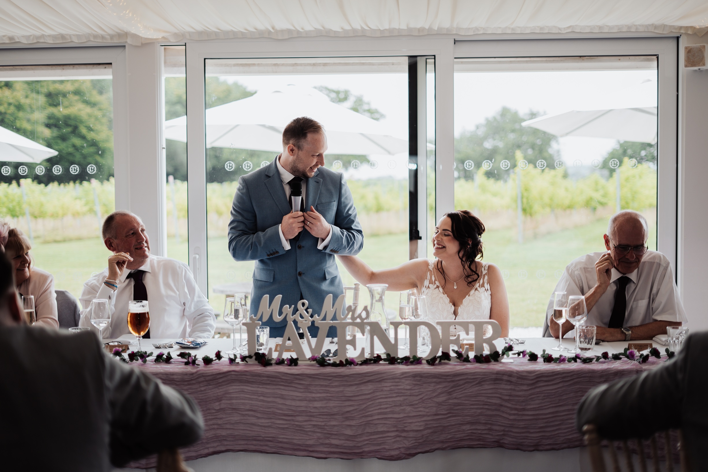 A candid  photo of a groom giving a speech or toast during the wedding reception. He holds a glass of 
                        champagne with a surprised, open-mouthed expression.
