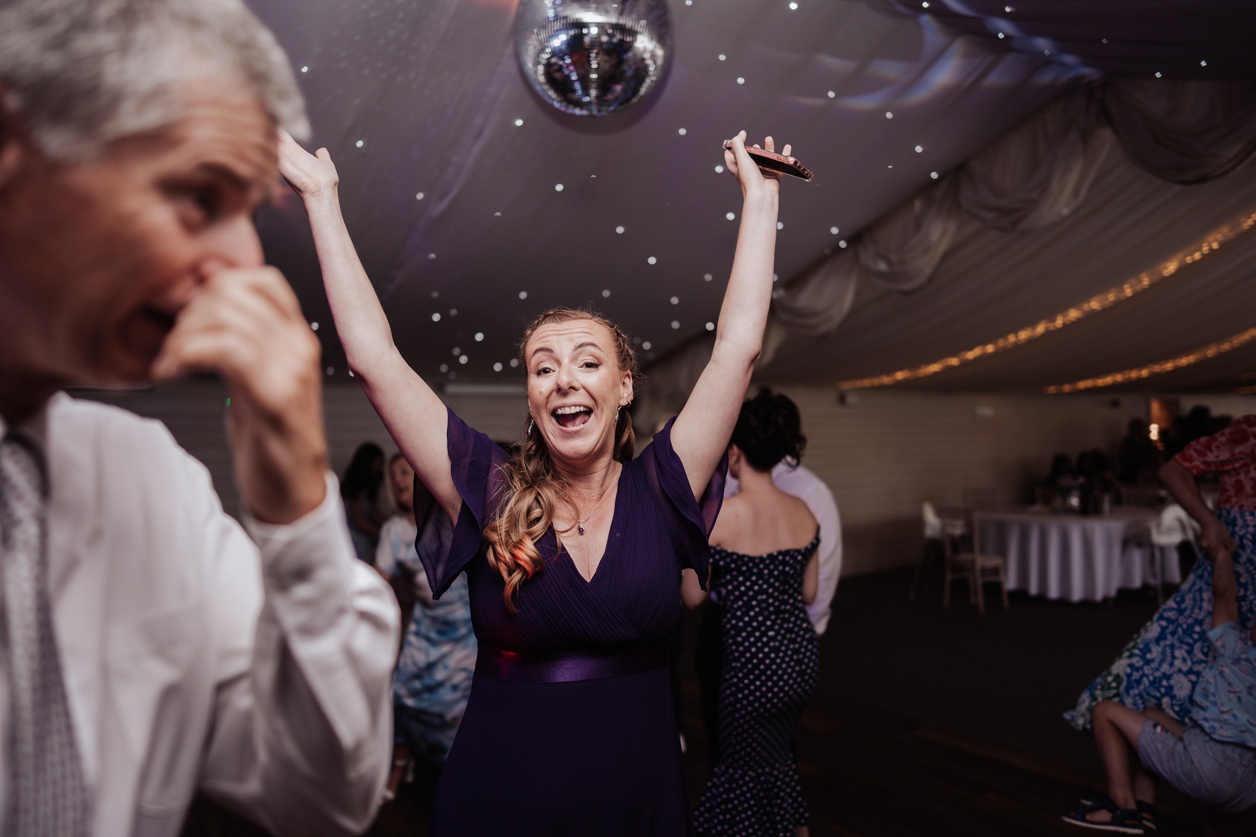 A wedding guest, wearing a dark purple dress, raises her hands in the air with a joyful, open-mouthed smile while 
                        dancing at the Llanerch Vineyard reception. A disco ball hangs from the ceiling.