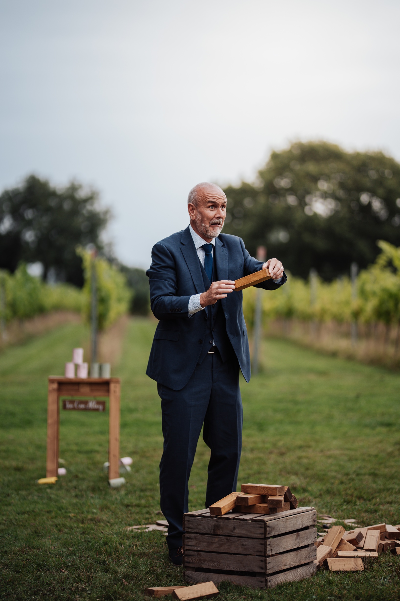 A man in a blue suit smiles while playing an outdoor lawn game, holding a wooden block from a Jenga tower. 
                        The photo captures a fun, candid moment from the wedding reception.