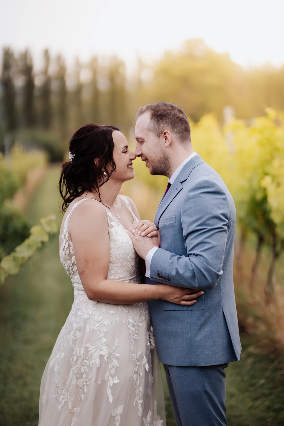 A portrait of a happy bride and groom standing in a vineyard at Llanerch Vineyard, with golden light in the background. The couple 
                                    is looking at each other, holding hands, and smiling.