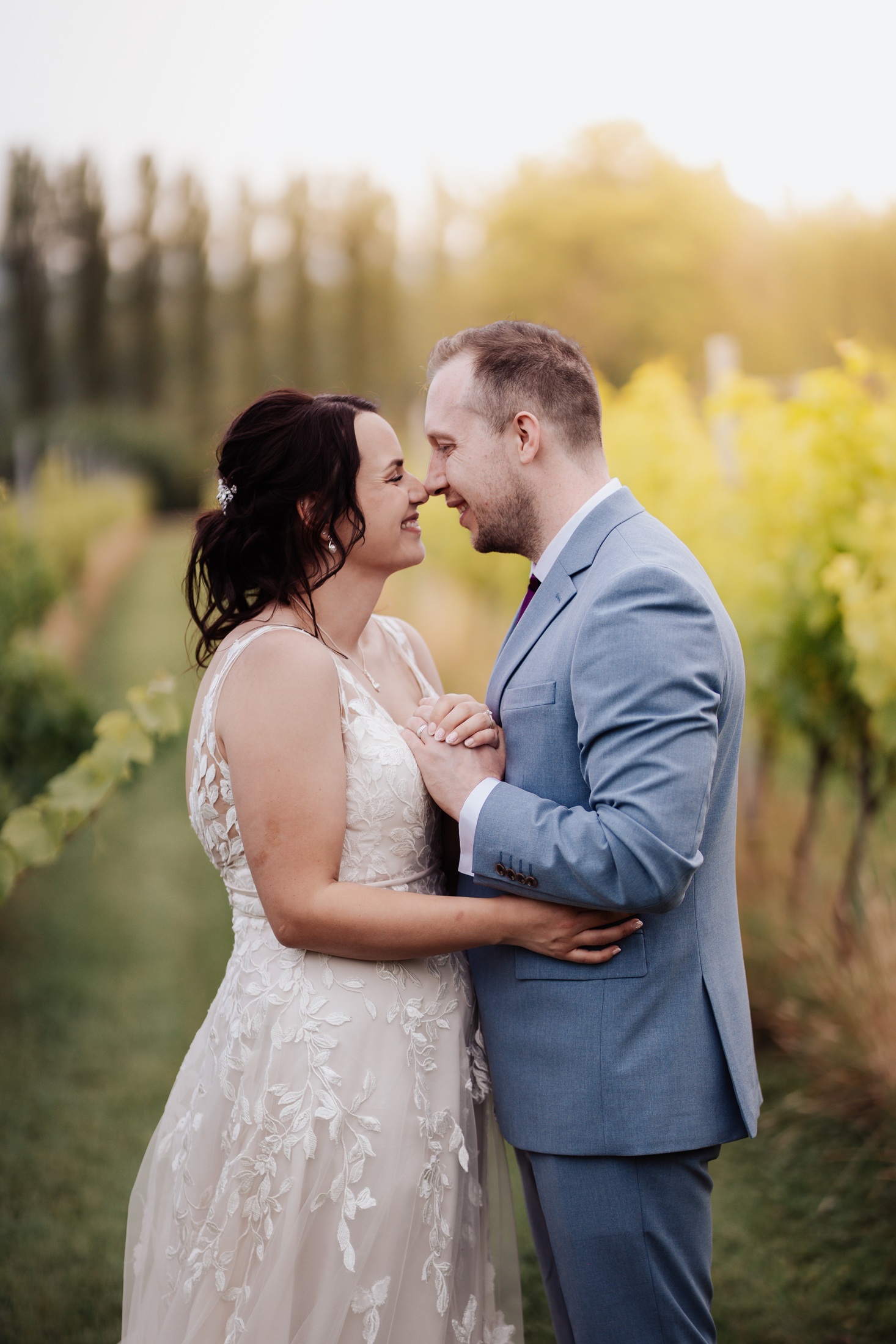 A portrait of a happy bride and groom standing in a vineyard at Llanerch Vineyard, with golden light in the background. The couple 
                        is looking at each other, holding hands, and smiling.