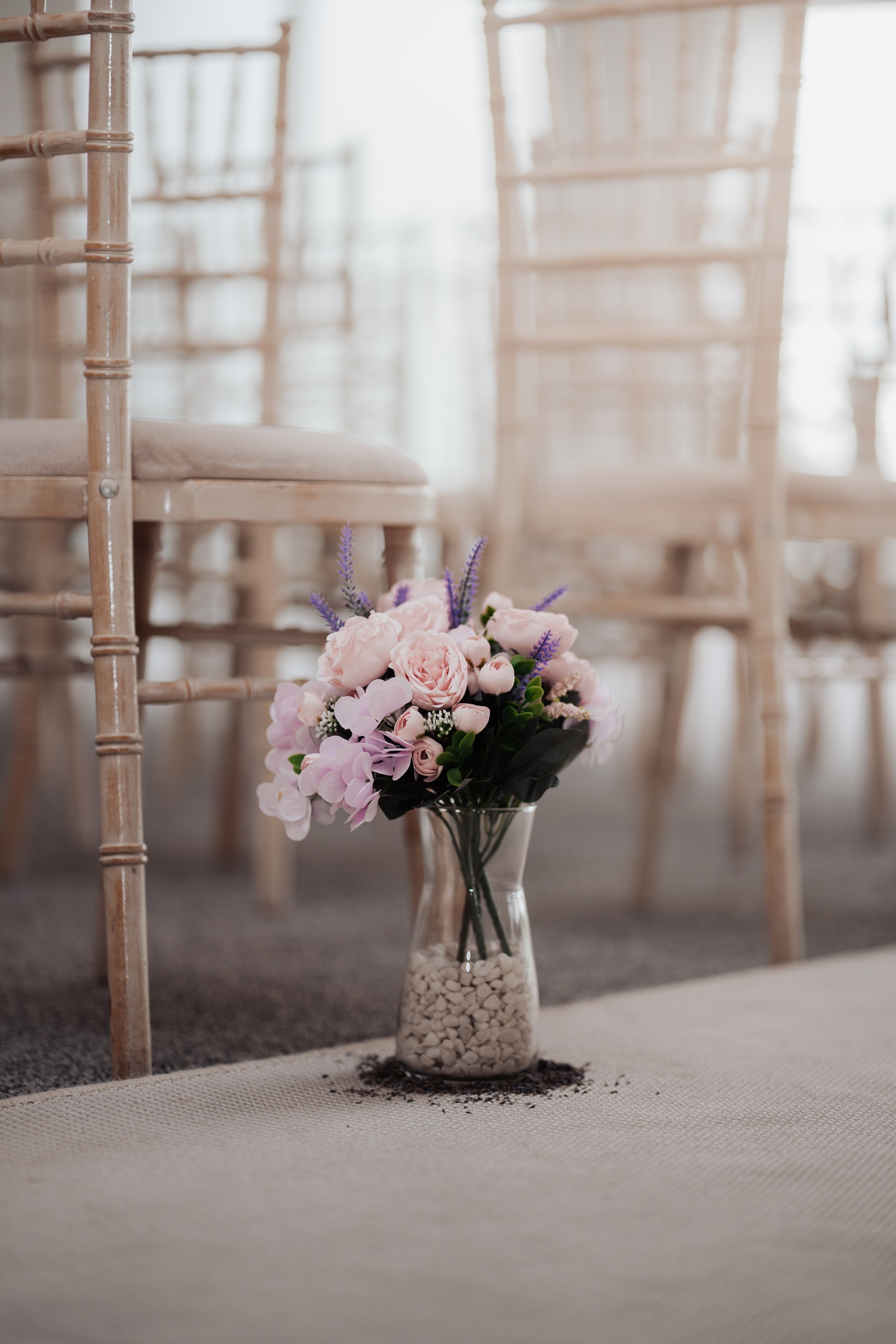 A close-up shot of the wedding ceremony decor, showing a floral arrangement in a glass vase placed on a carpeted aisle, with chairs and soft lighting in the background.