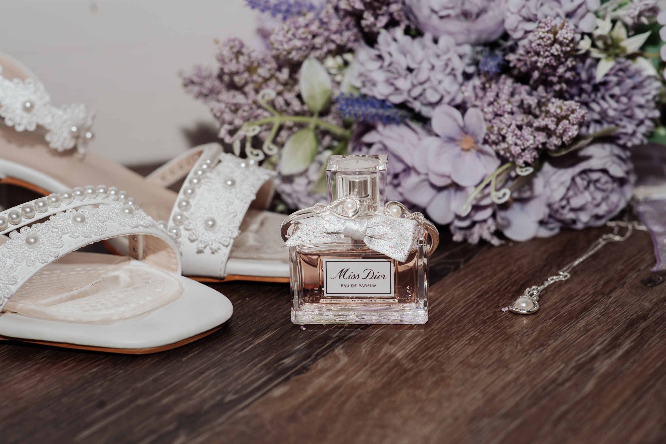 A flat-lay photo of a bride's wedding details, including a pair of white high-heeled shoes with pearls, a bottle of 
                        perfume, a necklace, and a floral bouquet.