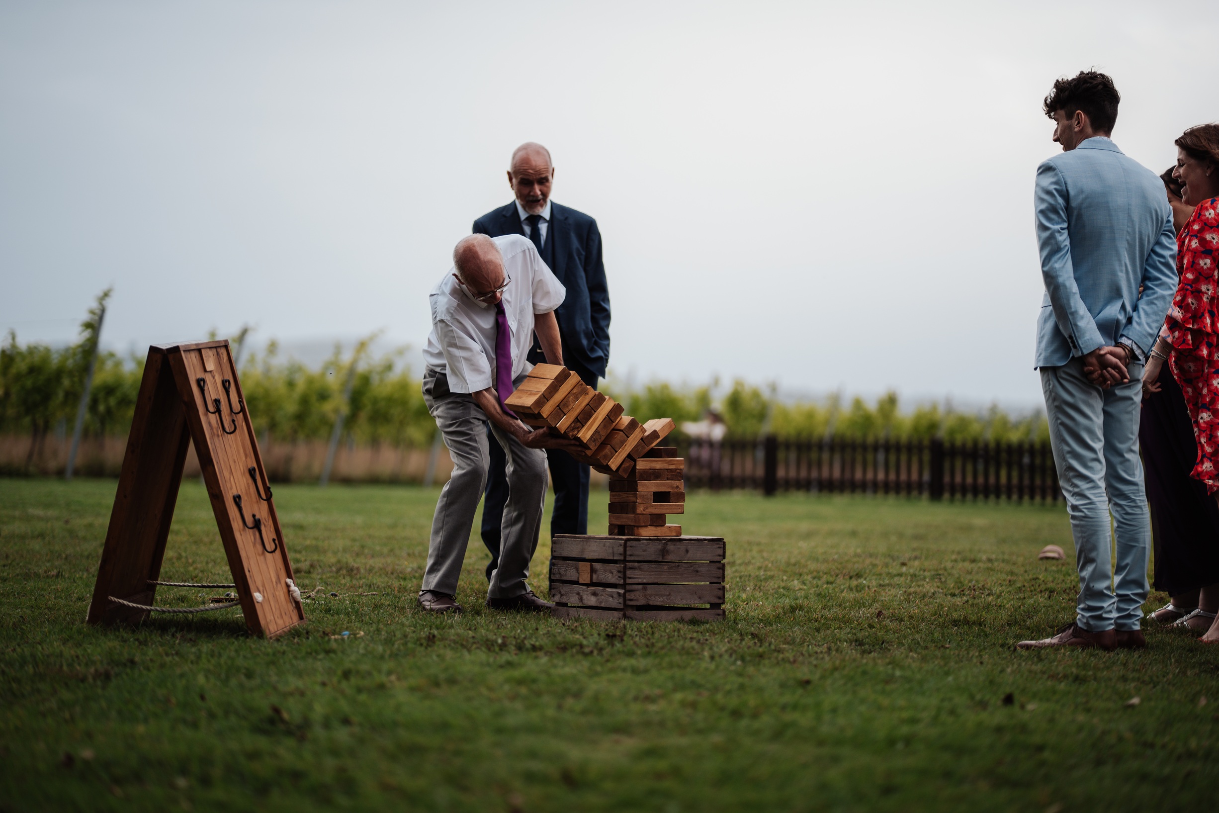 Wedding guests play a giant Jenga game on the lawn at Llanerch Vineyard in South Wales. The group is 
                        casually dressed and enjoying the outdoor reception.