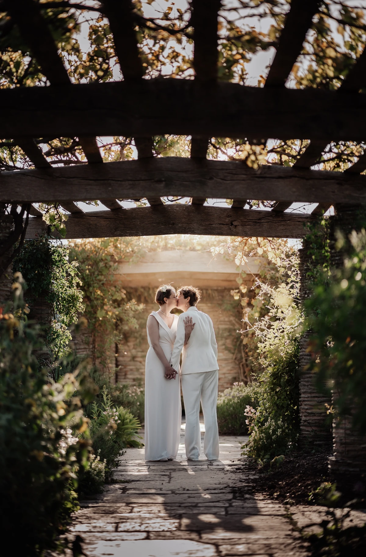 Romantic garden portrait of a same-sex couple sharing a kiss under a wooden pergola at Hestercombe Gardens in Taunton, bathed in golden hour sunlight.