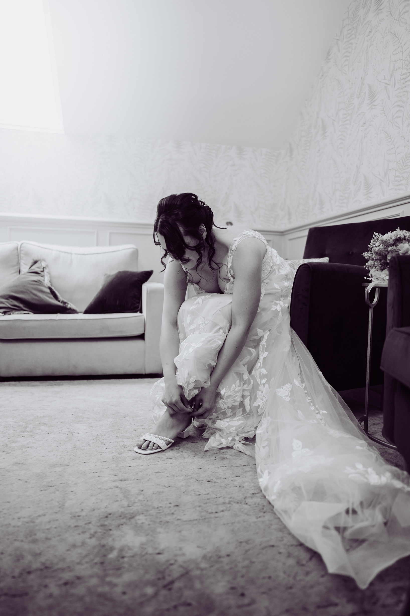 A black and white photo of a bride sitting on the floor, getting ready to put on her shoes. She is wearing a beautiful lace wedding dress 
                            and is in a room with a patterned wallpaper and a couch.